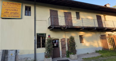 Two-story yellow trattoria with a long wooden balcony, doorway, and potted shrubs at the entrance.