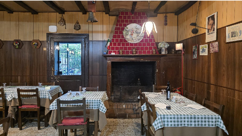 Cozy, wood-paneled dining room with checkered tablecloths, four tables set for dining, and a brick fireplace as a focal point.