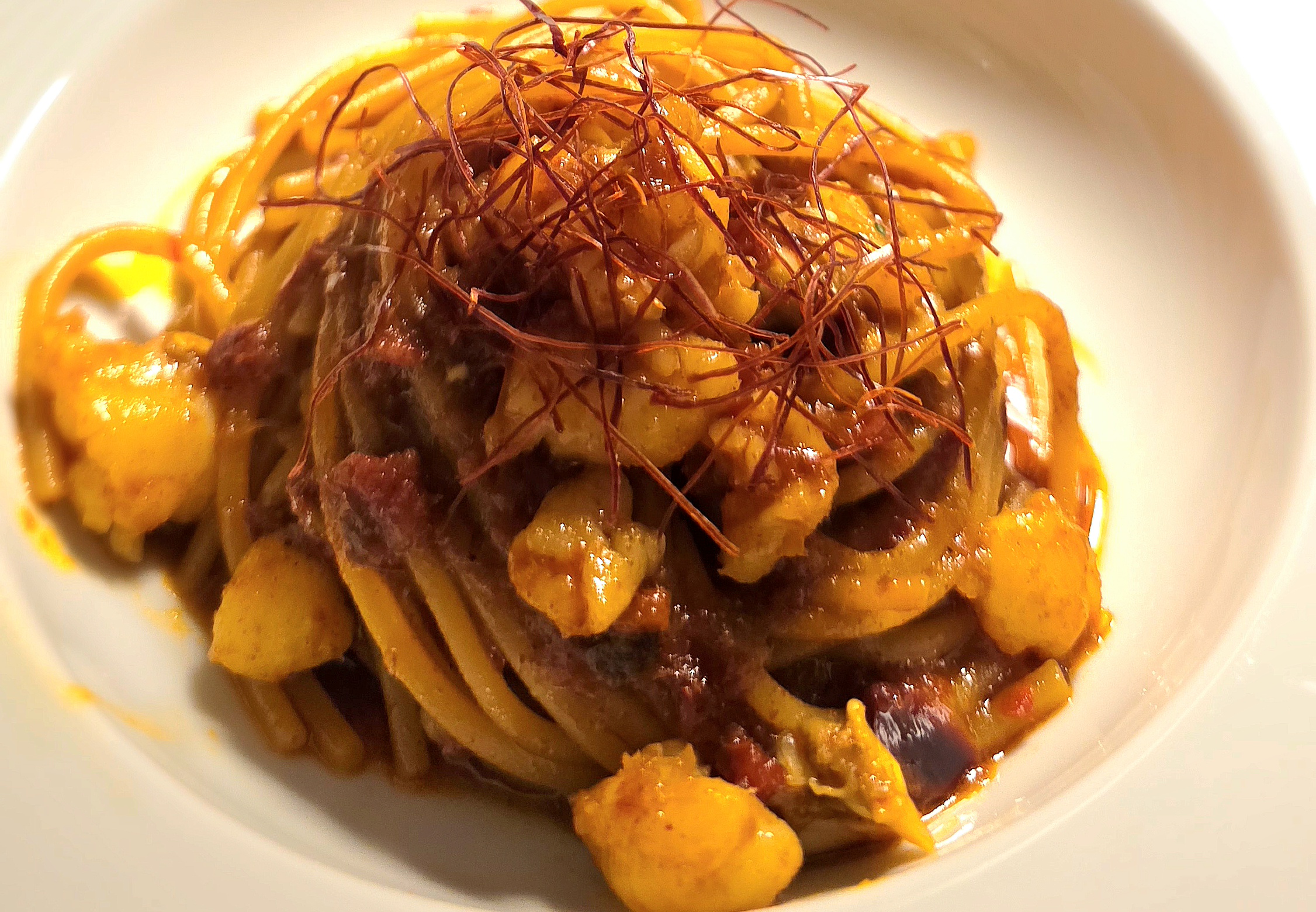 Pasta in a white bowl with tomato sauce, sausage chunks, shrimp, and a tangle of thin red garnish on top.