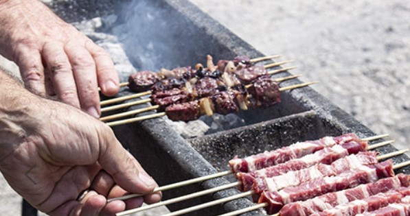 Hands thread meat onto wooden skewers above a charcoal grill, ready to barbecue. The skewers include chunks of beef cooking over open heat.
