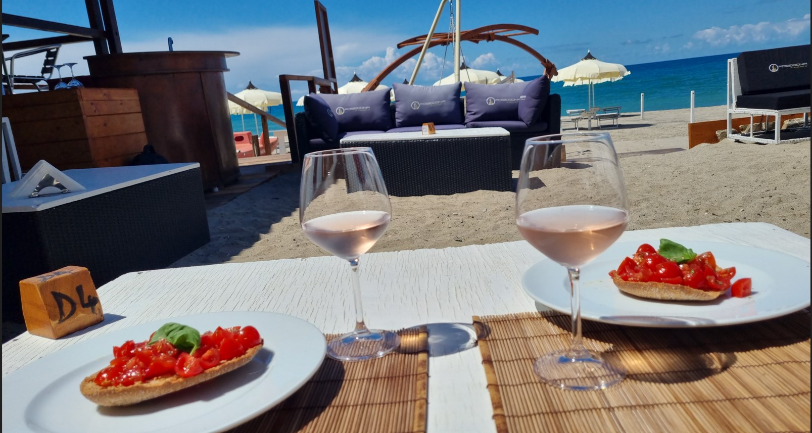 Beachclub_poseidonia_cilento_colzaione | Food and Wine Magazine Beachfront dining setup with two plates of tomato bruschetta and two glasses of rosé on a white table, sandy beach and blue sea in the background.