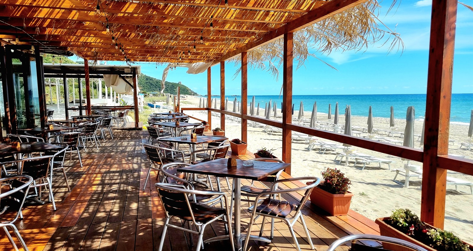 Beachclub_poseidonia_cilento_copertina | Food and Wine Magazine Outdoor beachside cafe on a wooden deck with straw roof, metal chairs and tables overlooking a sandy shore and blue sea.