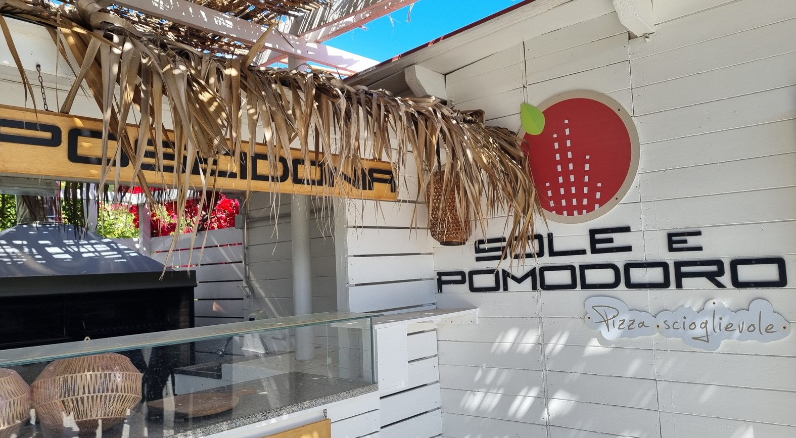 Beachclub_poseidonia_cilento_pizzeria | Food and Wine Magazine Outdoor pizza counter with a thatched roof, white wooden wall, and a round red logo; wicker baskets sit in front of a glass display case.