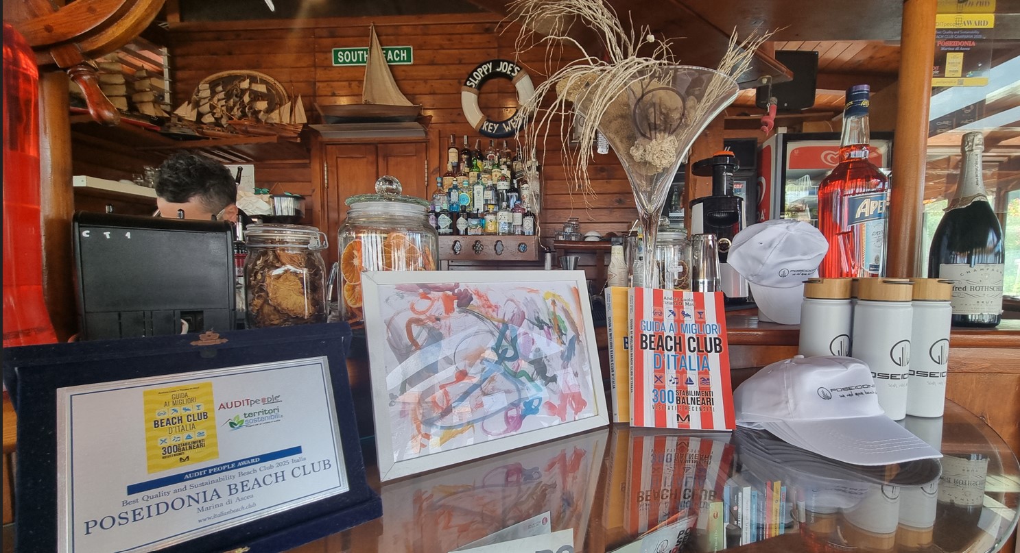 Beachclub_poseidonia_cilento_premi | Food and Wine Magazine Bar counter scene with framed award certificates, a colorful abstract art print, white caps, and bottles on a shelf behind.