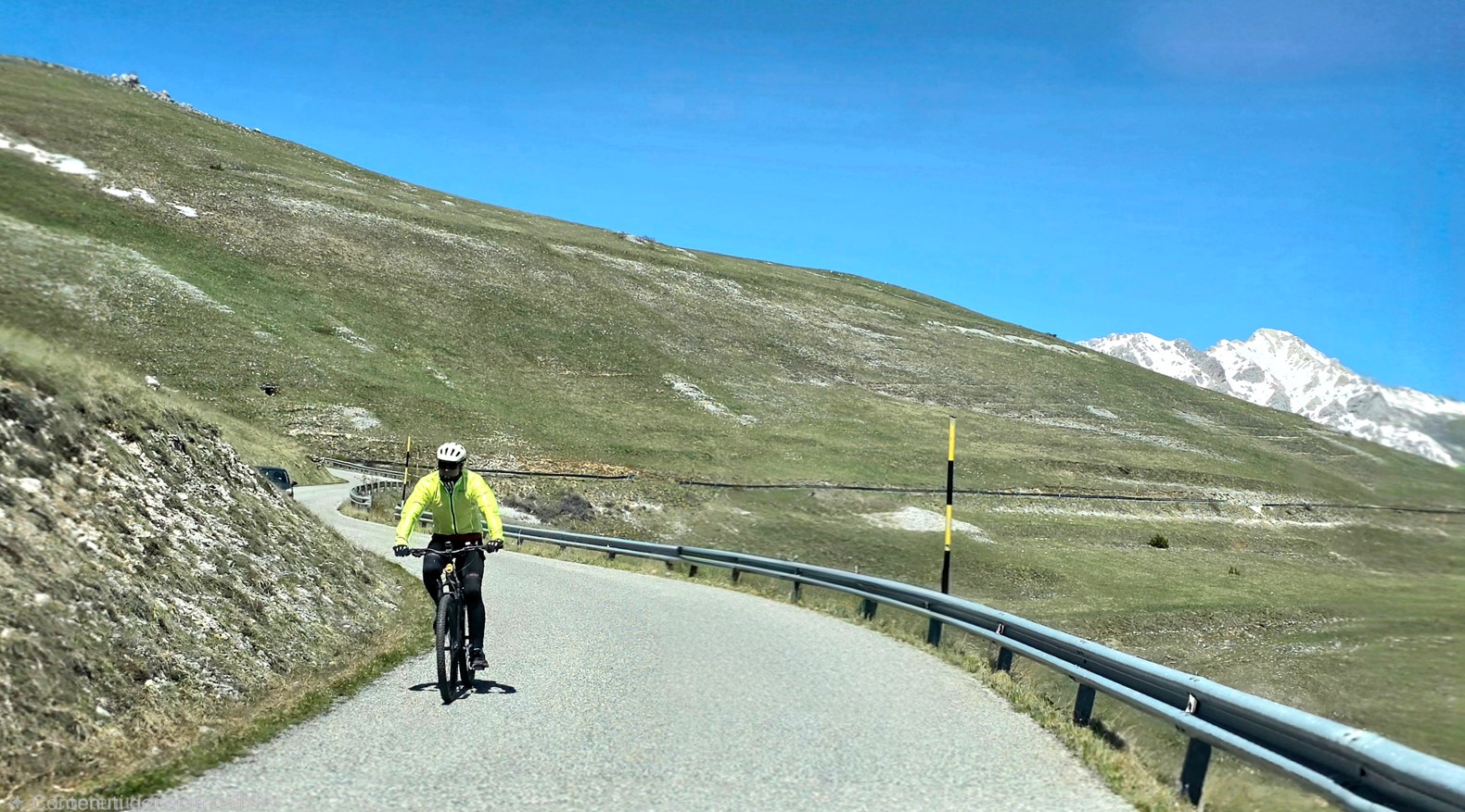 Cyclist wearing a neon jacket rides along a winding mountain road with a metal guardrail and grassy slopes, snow-capped peaks in the distance under a clear blue sky.