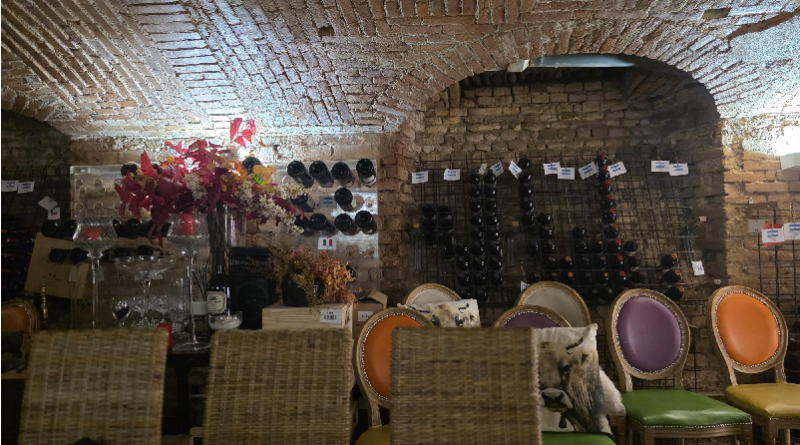 Wine tasting room in a brick cellar with bottles on wall racks, a table set with glassware and flowers, and colorful chairs along the side.