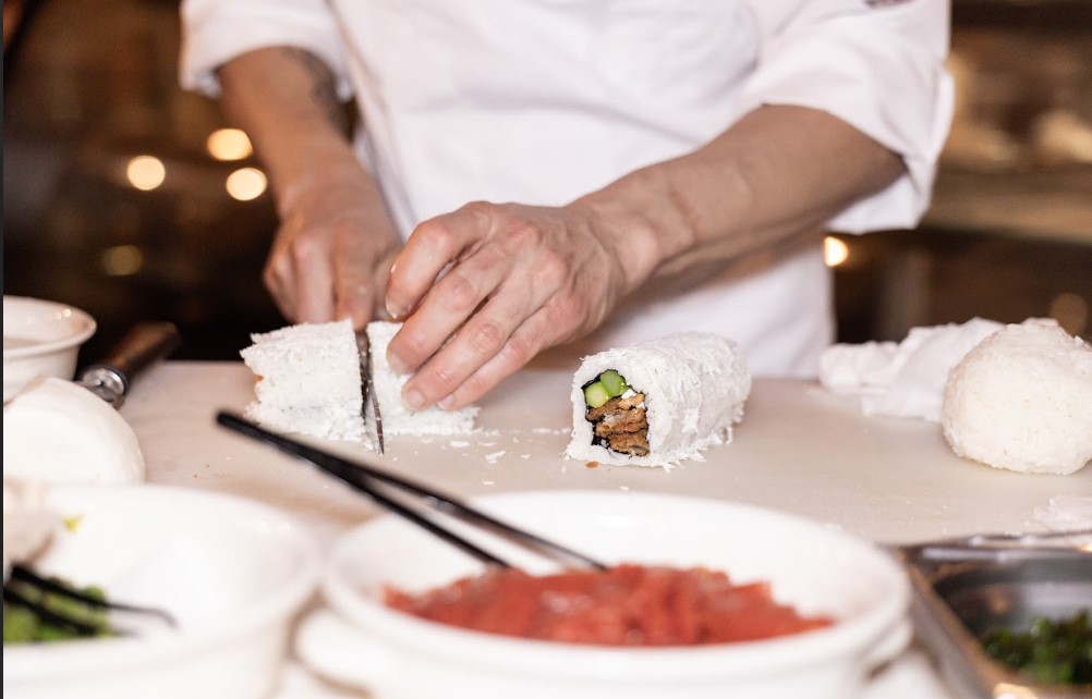 Cilento_roll | Food and Wine Magazine Chef cutting white-rice sushi rolls on a cutting board; one roll sliced open shows cucumber inside the filling