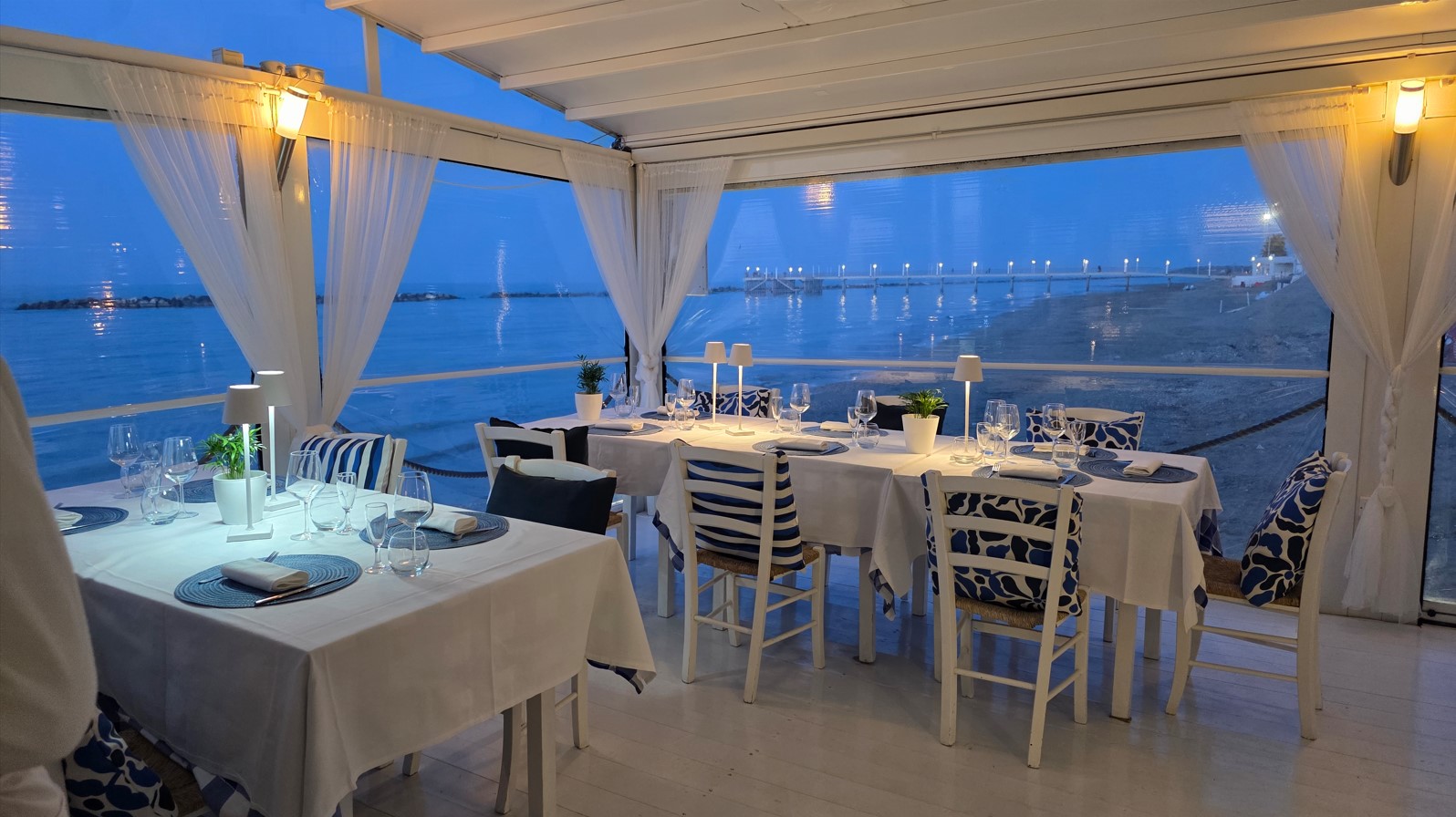 Seaside restaurant dining area with white tables, blue cushions, and sheer curtains, overlooking the water at dusk.