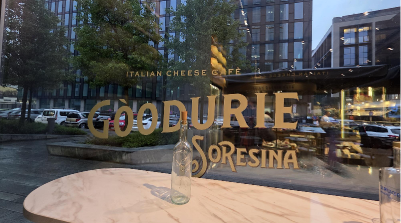Storefront window displaying signage for an Italian cheese cafe; text on the glass reads 'Italian Cheese Café' and 'GOODURIE SORESINA'; a glass bottle sits on a round marble table outside, with reflections of the street and parked cars.