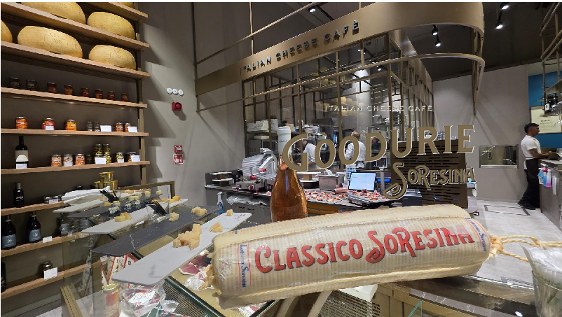 Goodurie Soresina_interni | Food and Wine Magazine Glass display of cheeses at an Italian cheese cafe, with a large wrapped cheese log labeled 'Classico' in the foreground and shelves of jars in the background.