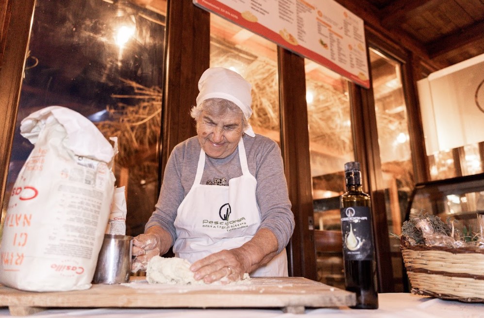 Nonna_ines | Food and Wine Magazine Older woman in a white apron and headscarf kneading dough on a wooden table in a cozy kitchen; flour and a bag of flour nearby.
