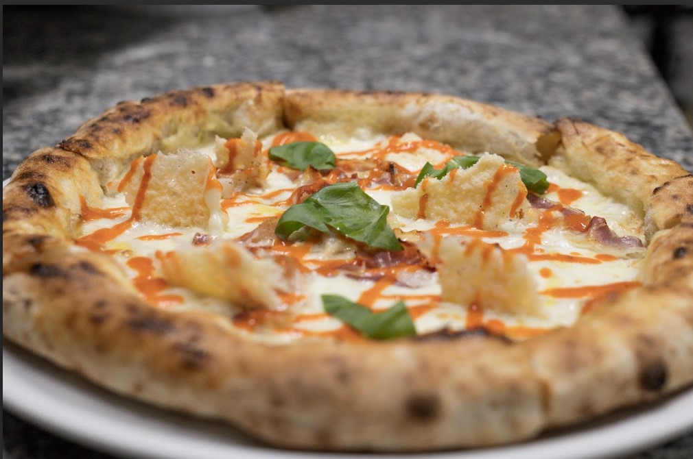 Pizza_gourme_poseidonia | Food and Wine Magazine Wood-fired pizza topped with mozzarella, pineapple chunks, bacon bits, fresh basil, and an orange drizzle on a blistered crust