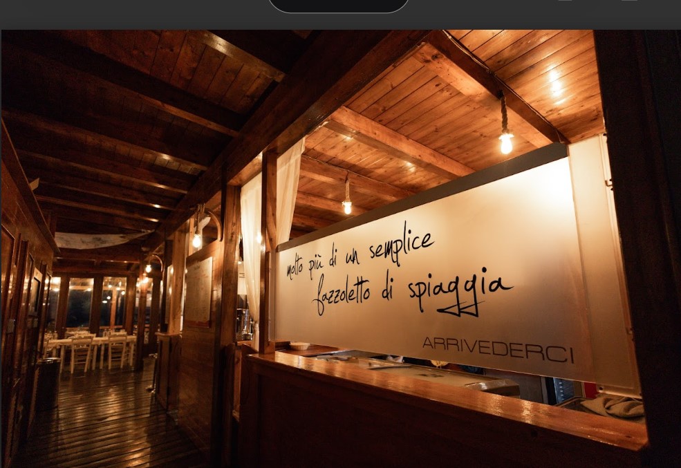 Poseidonia_by_night | Food and Wine Magazine Cozy wooden restaurant interior at night with warm lighting and a frosted sign displaying Italian text, including 'ARRIVEDERCI'