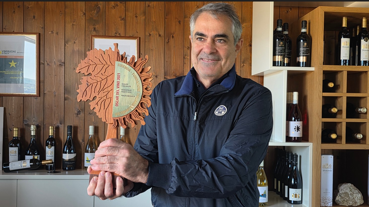 Man in a navy jacket holding a round wooden award plaque in a wine shop with bottles on shelves behind him.