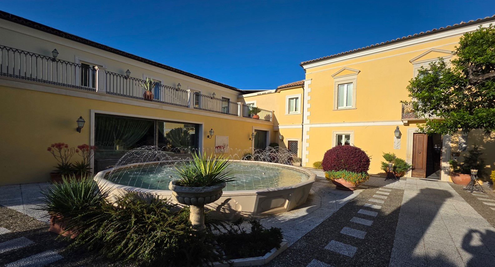 Yellow Mediterranean courtyard with a circular fountain and potted plants in front of a two-story building under a blue sky.