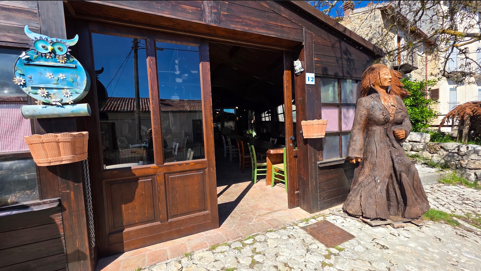 Open wooden storefront with glass doors, a blue owl mailbox on the left, hanging baskets, and a carved wooden woman statue outside on a cobblestone path