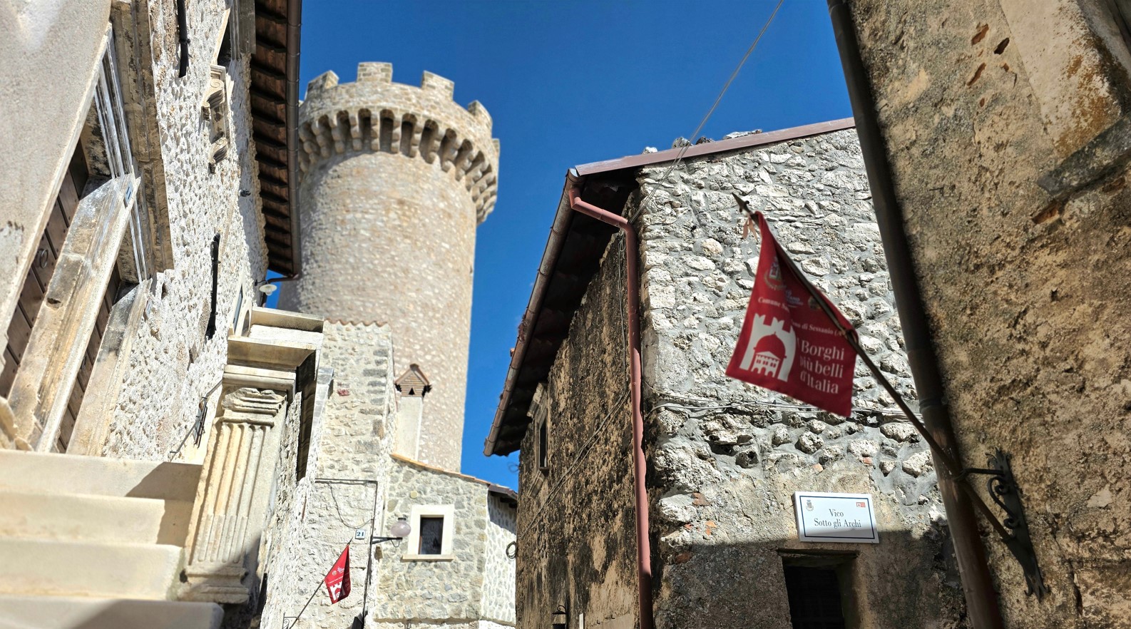 Narrow stone street in a historic Italian village, with a round crenellated tower on the left and blue sky above, red banners visible