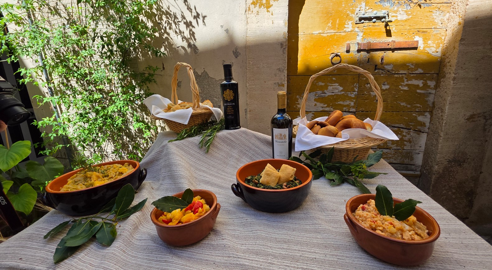 Outdoor table setting with four clay bowls of prepared dishes, a basket of bread, and two wine bottles against a sunlit, rustic wall backdrop.