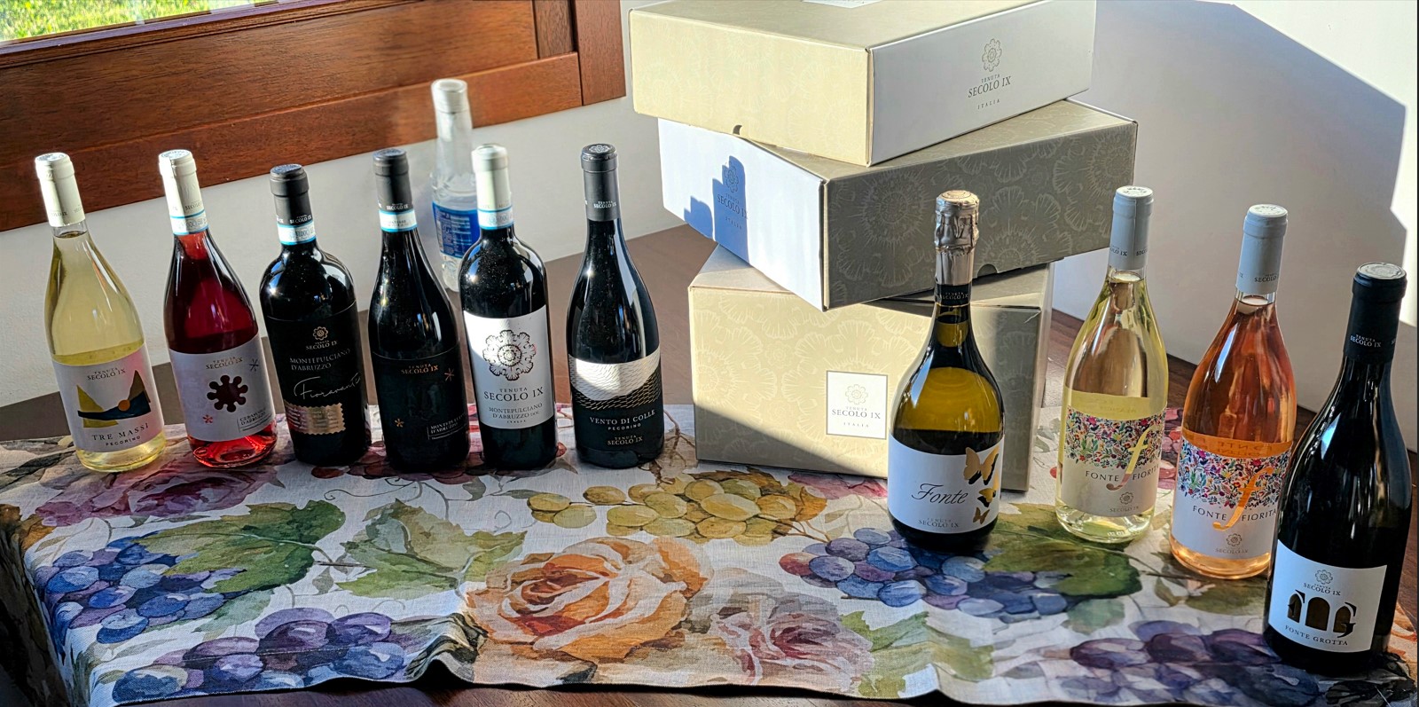 Row of wine bottles in varying colors (white, red, rosé) lined up on a floral tablecloth with beige gift boxes stacked in the background.
