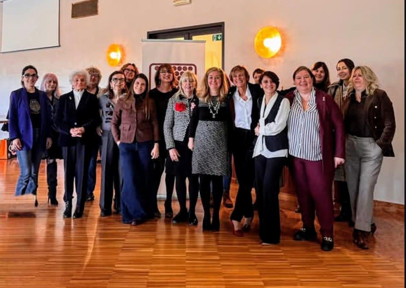 Donne_del_vino_della_lombardia | Food and Wine Magazine Group of about 15 women posing for a group photo in a bright conference room with a wood floor and a banner in the background.