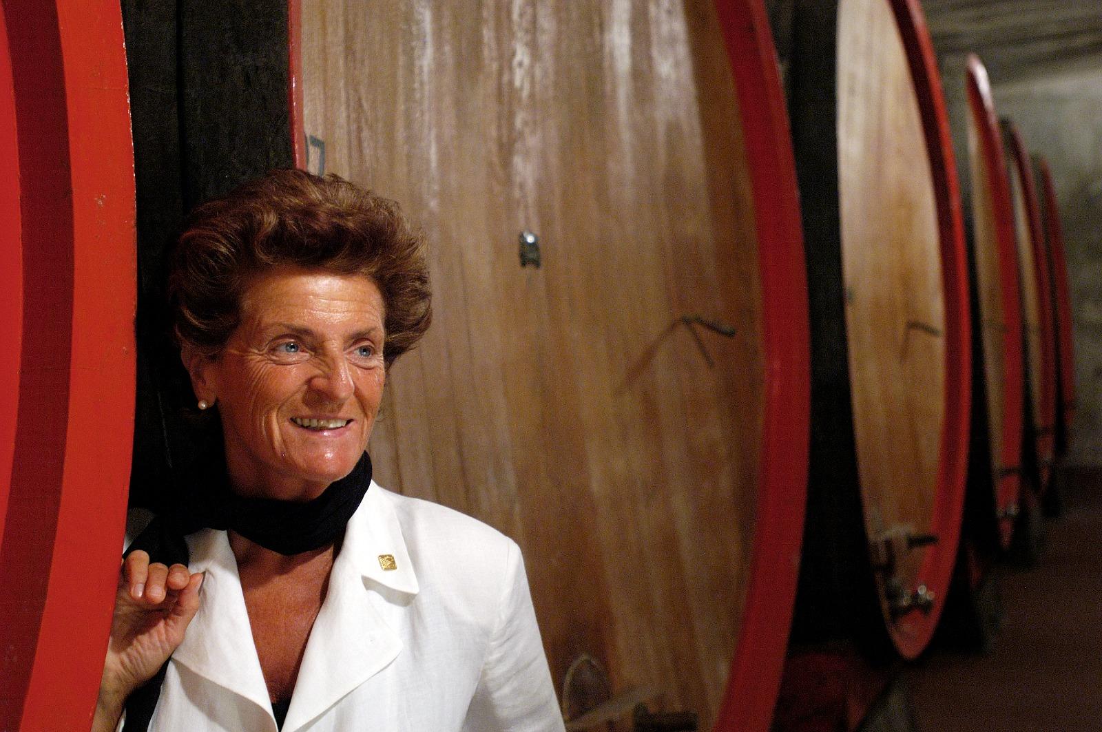 Pia_berlucchi | Food and Wine Magazine Smiling woman in a white blazer and black scarf standing next to large wooden wine barrels in a cellar.