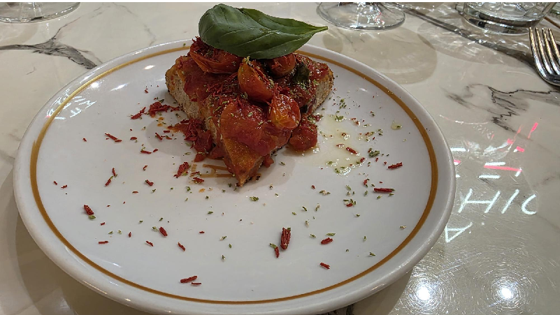 Tomato-based crostini stack topped with a basil leaf on a white plate, garnished with scattered herbs.