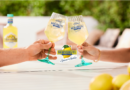 Two people toast with pale yellow wine glasses labeled Benvenuti, outdoors on a sunny table with lemons nearby.