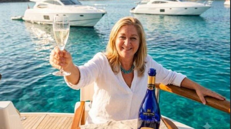 Woman on a yacht toasting with a flute of champagne, turquoise water and boats in the background.