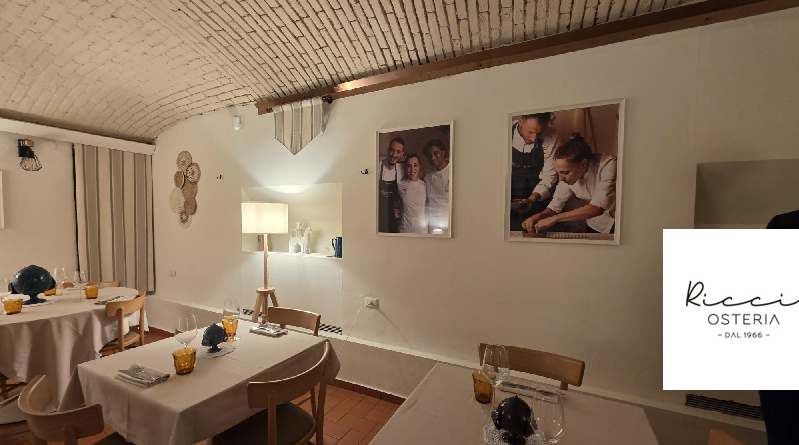 Ricciosteria_sala Interni | Food and Wine Magazine Cozy dining room with white-clothed tables, warm lighting, and framed chef photos on a curved ceiling wall. A logo appears in the corner.