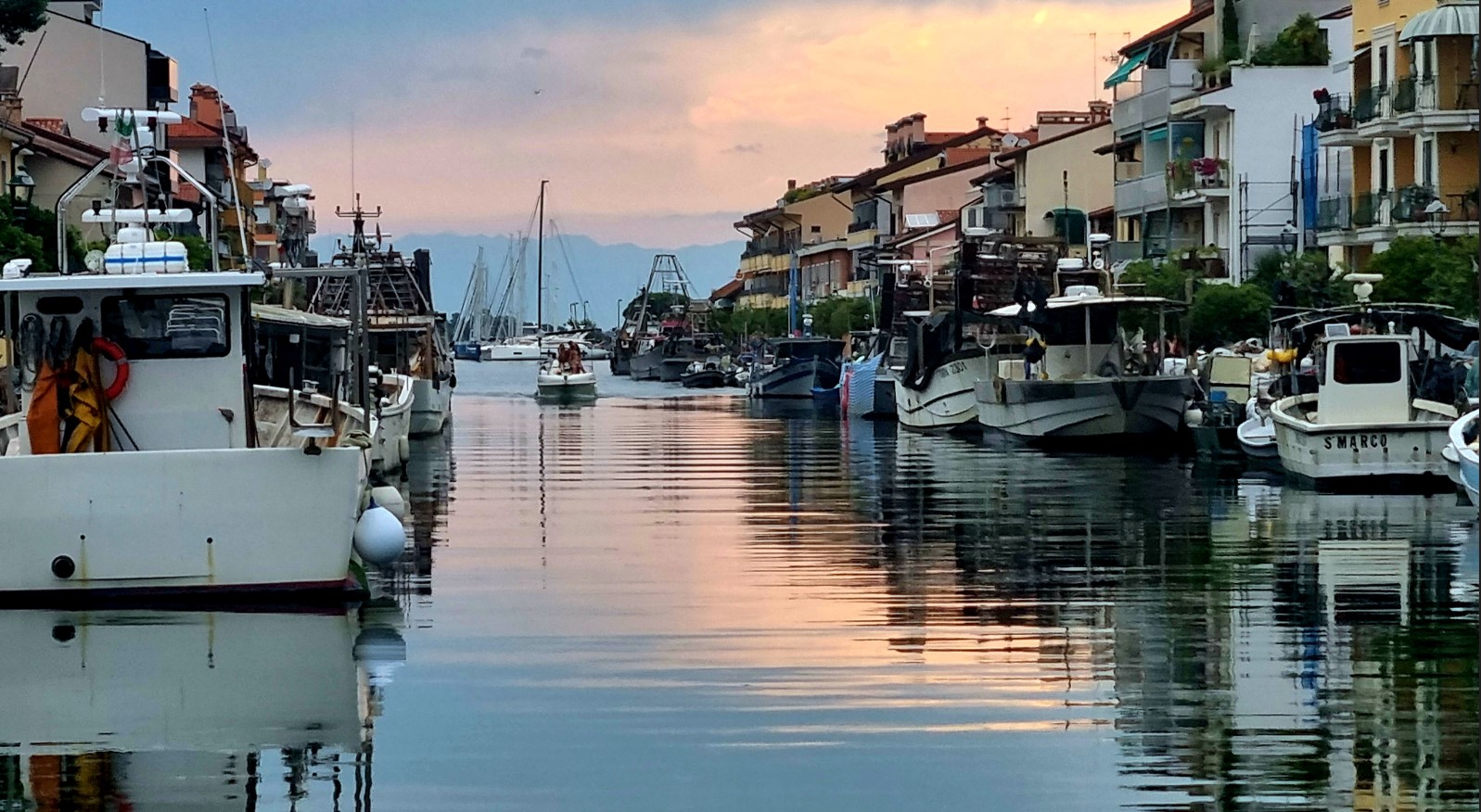 Sunset over a narrow harbor canal lined with boats and colorful buildings, reflections ripple on the water's surface.