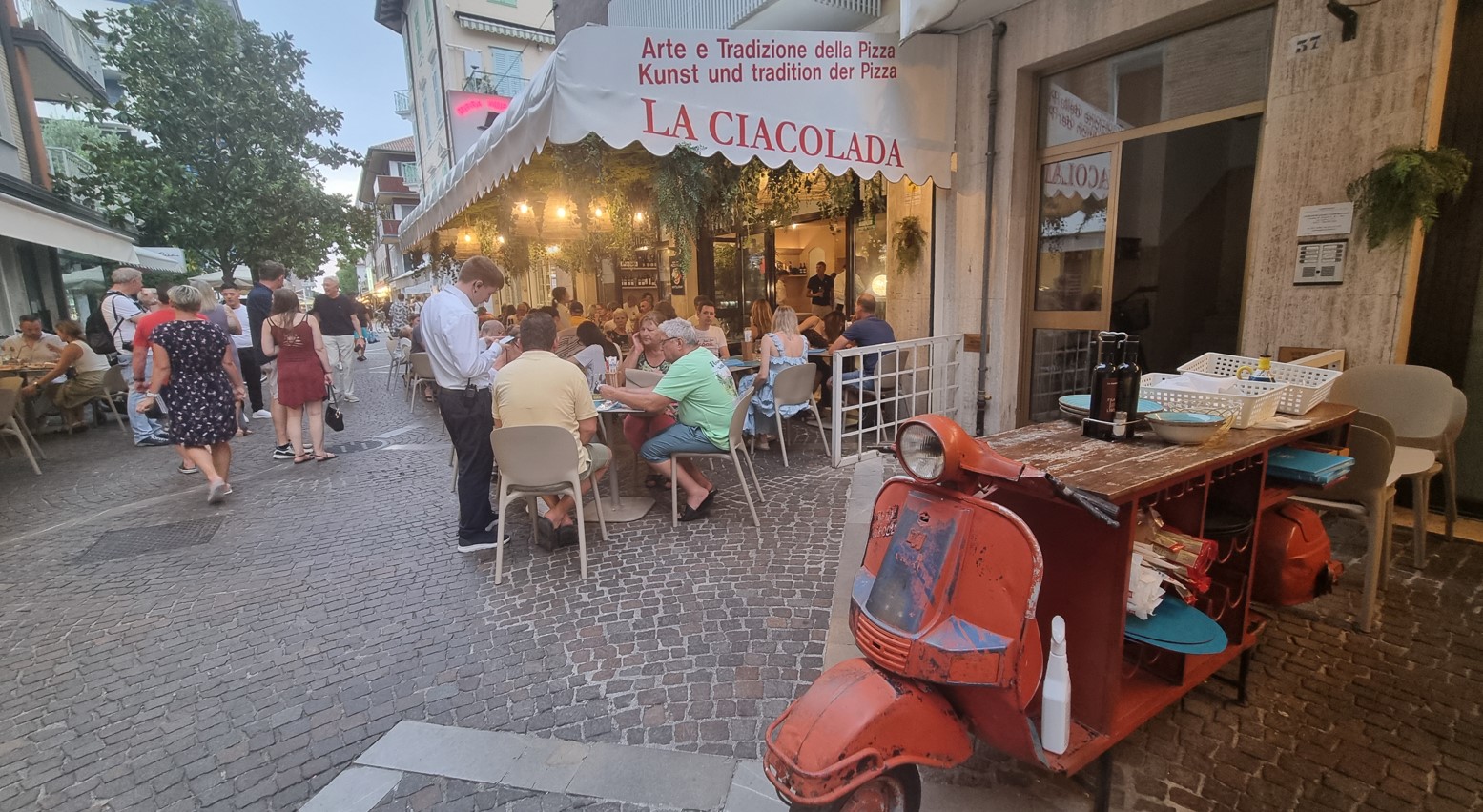 Outdoor restaurant on a cobblestone street with diners under a white awning; a red vintage scooter sits at the foreground side of the scene.