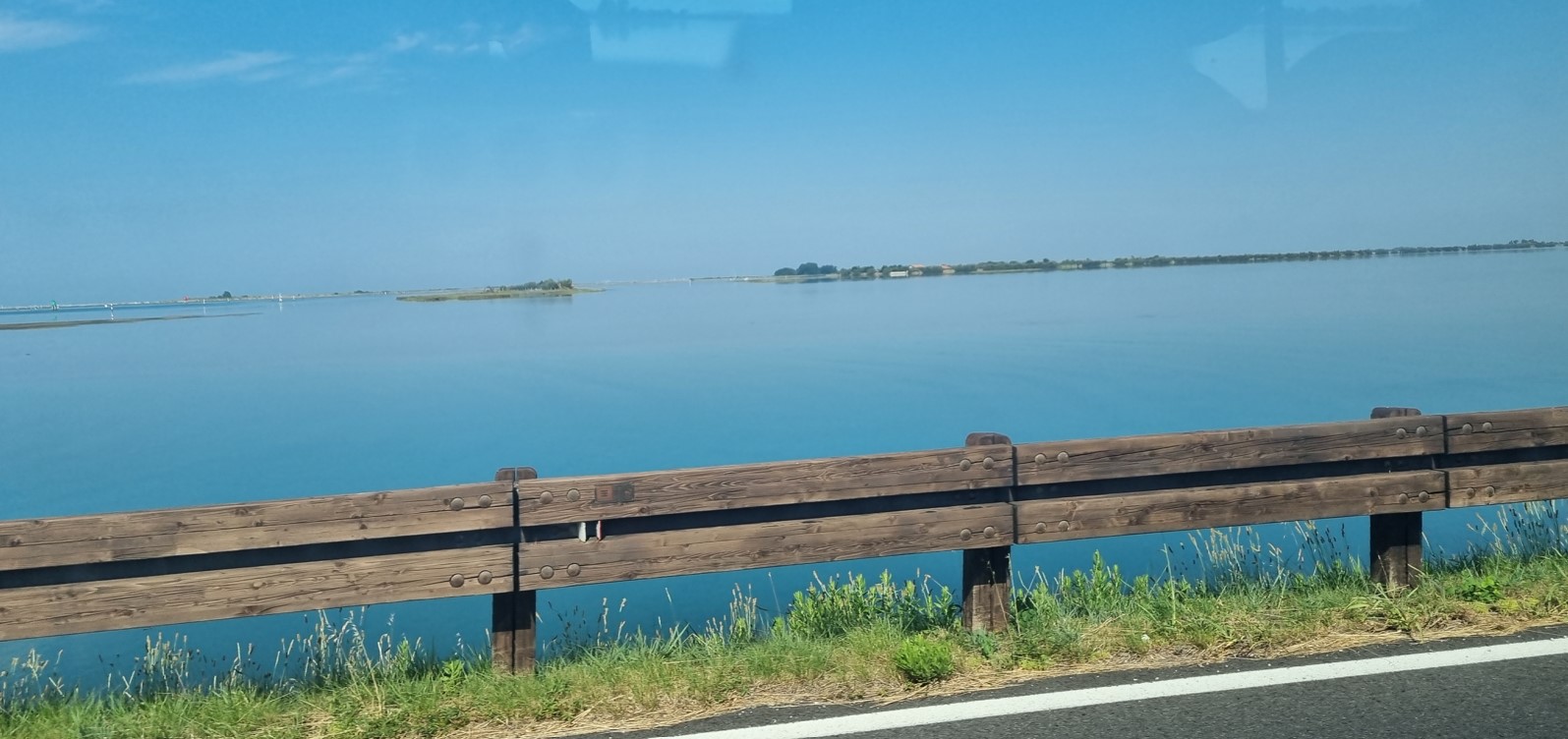 Blue lake seen from a roadside, with a weathered wooden guardrail in the foreground and distant small islands on the water.