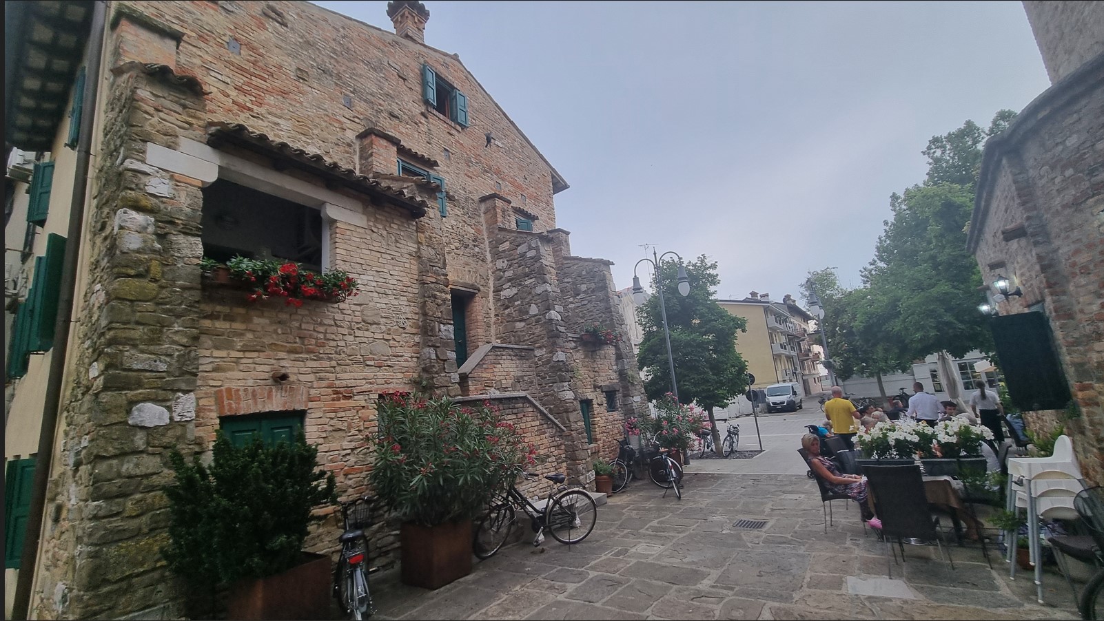 Stone building with flower boxes on a cobblestone street; bikes parked outside and a cafe with outdoor seating.