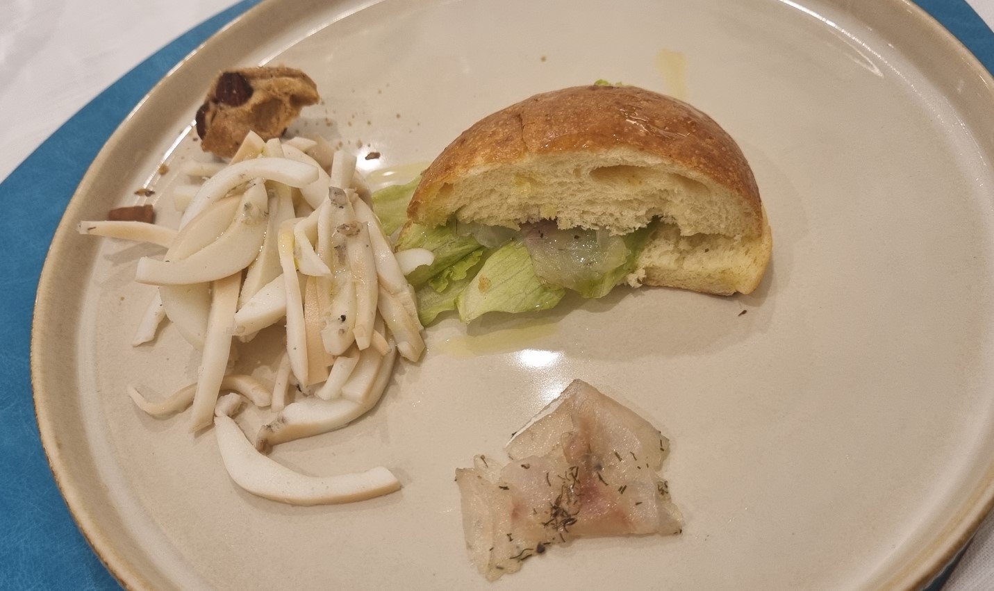 La Ciacolada_cucina_pesce | Food and Wine Magazine Plate with a half brioche bun sandwich filled with lettuce (and onion) on a beige plate, next to shredded white mushrooms and a small herb-seasoned fish fillet, plus a tiny cookie in the corner.