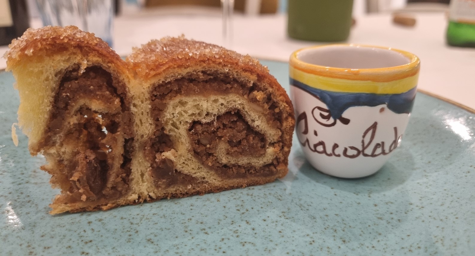 A sliced cinnamon roll pastry on a blue speckled plate next to a colorful ceramic mug with yellow rim.