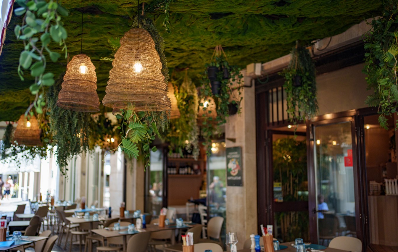 La Ciacolada_esternijpg | Food and Wine Magazine Outdoor cafe with wicker pendant lamps and a green leafy ceiling above dining tables.