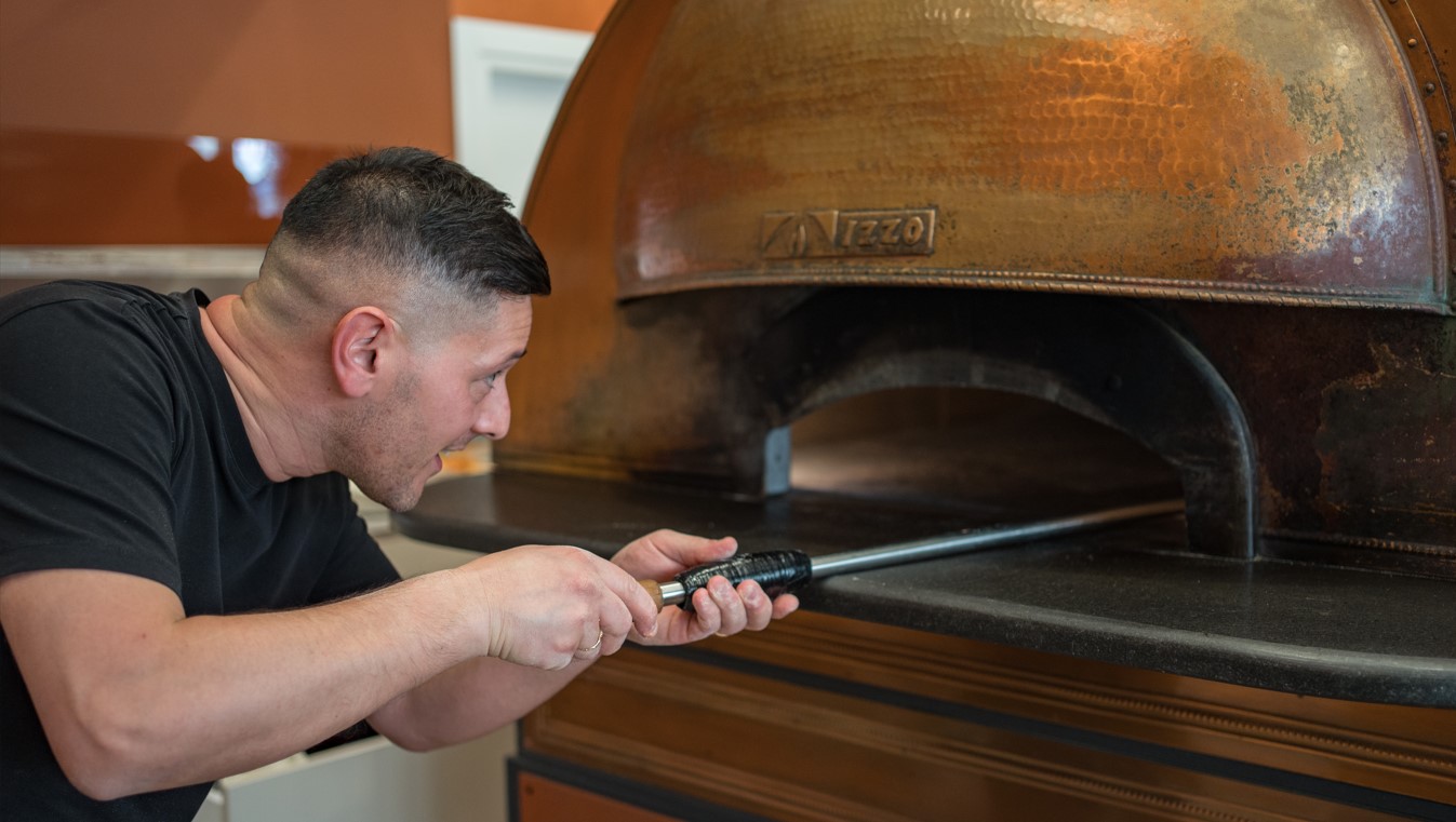 La Ciacolada_pizzaiolojpg | Food and Wine Magazine Man in a black T-shirt uses a long-handled tool to work on a large copper wood-fired pizza oven.