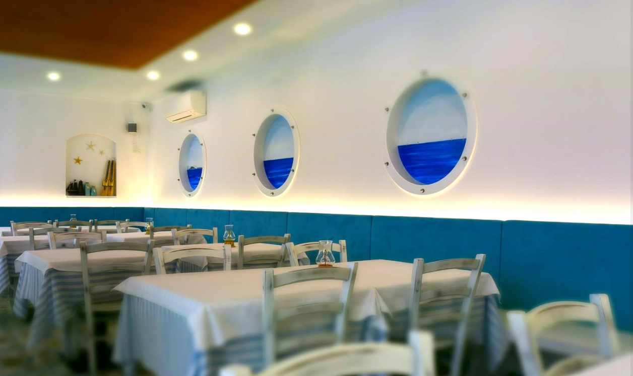 La_ciacolada_pizza_sale_interne | Food and Wine Magazine Seaside-themed dining room with white tables, white chairs, and blue bench seating along a wall, lit softly.