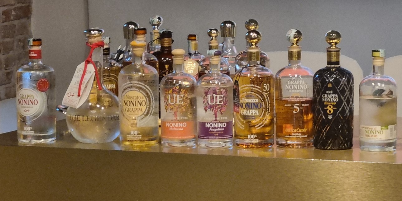 Row of assorted liquor bottles on a bar counter, mostly Grappa and Nonino labels, with a brick wall in the background.