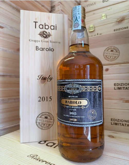 Bottle of Tabai Barolo Grappa, 2015, with a black-and-gold label, beside a wooden box engraved 'Barolo Italy 2015' in a crate-filled background.