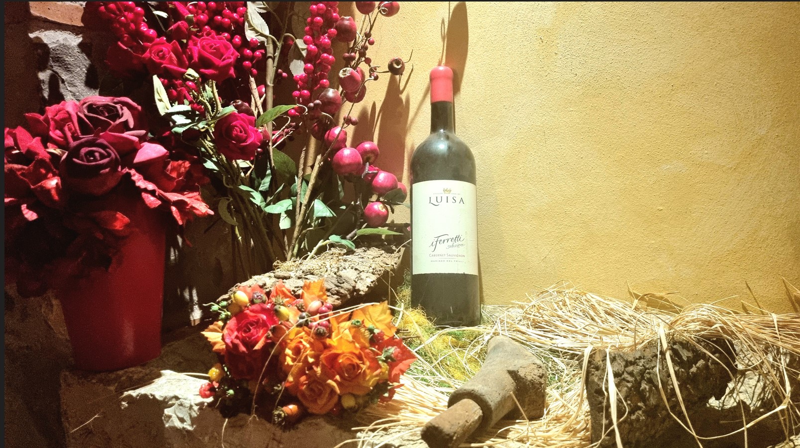 Bottle of Luisa Cabernet Sauvignon beside red rose bouquets and berry stems on a rustic stone ledge with straw against a yellow wall, still-life scene.