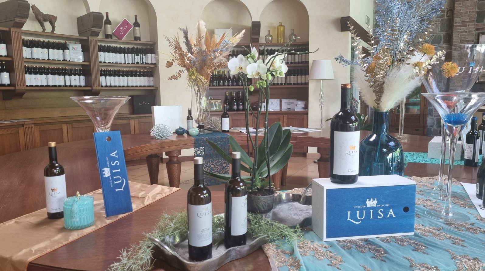 Wine display on a wooden table with blue Luisa branding, bottles, vases, and floral arrangements in a tasting room.