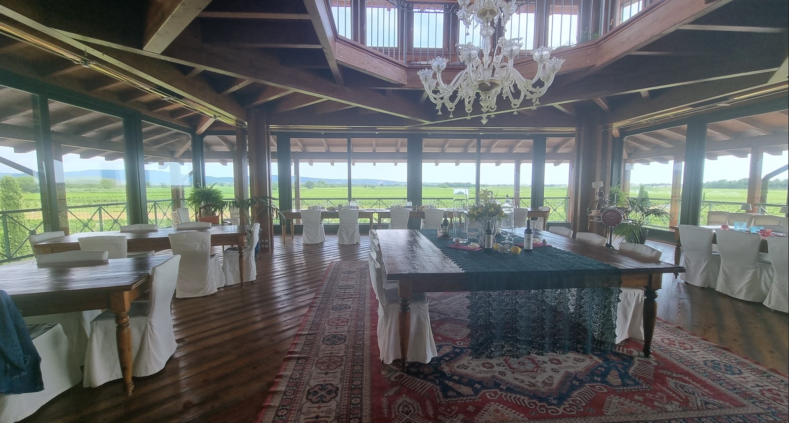 Rustic banquet hall with wooden beams, a crystal chandelier, and floor-to-ceiling windows overlooking green fields; a long central table with a blue lace cloth.